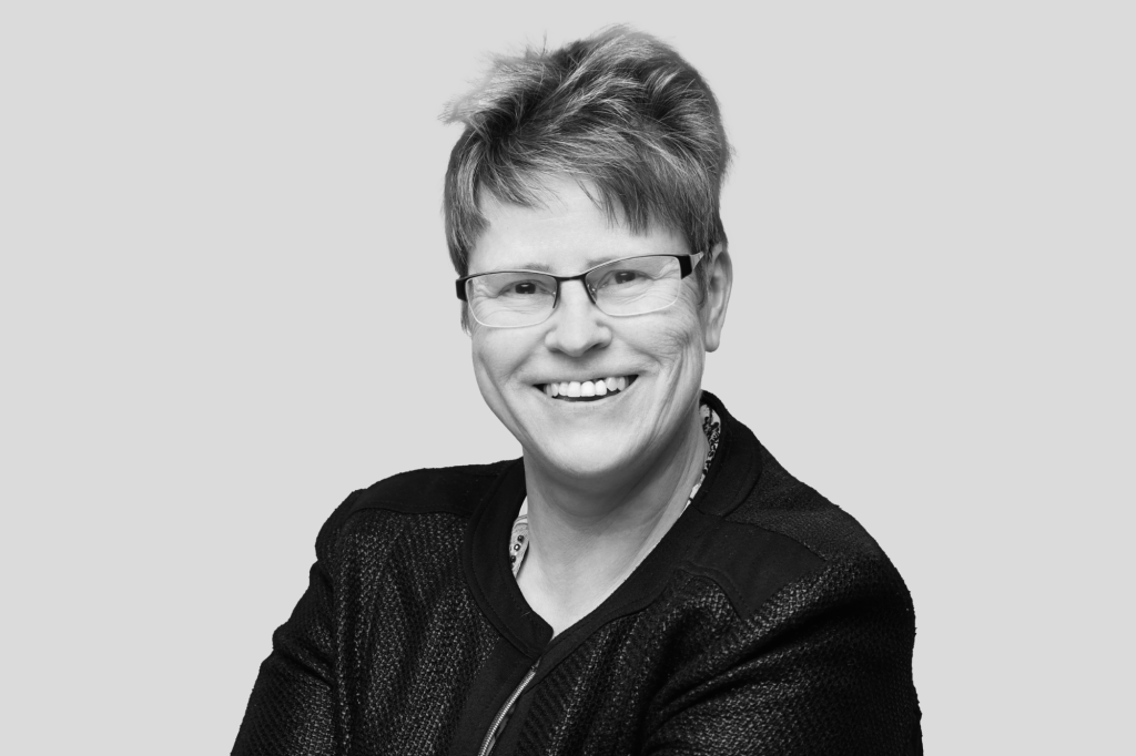 Schwarz-weiß Porträt einer Frau mit Brille und kurzer Frisur, die freundlich in die Kamera lächelt.