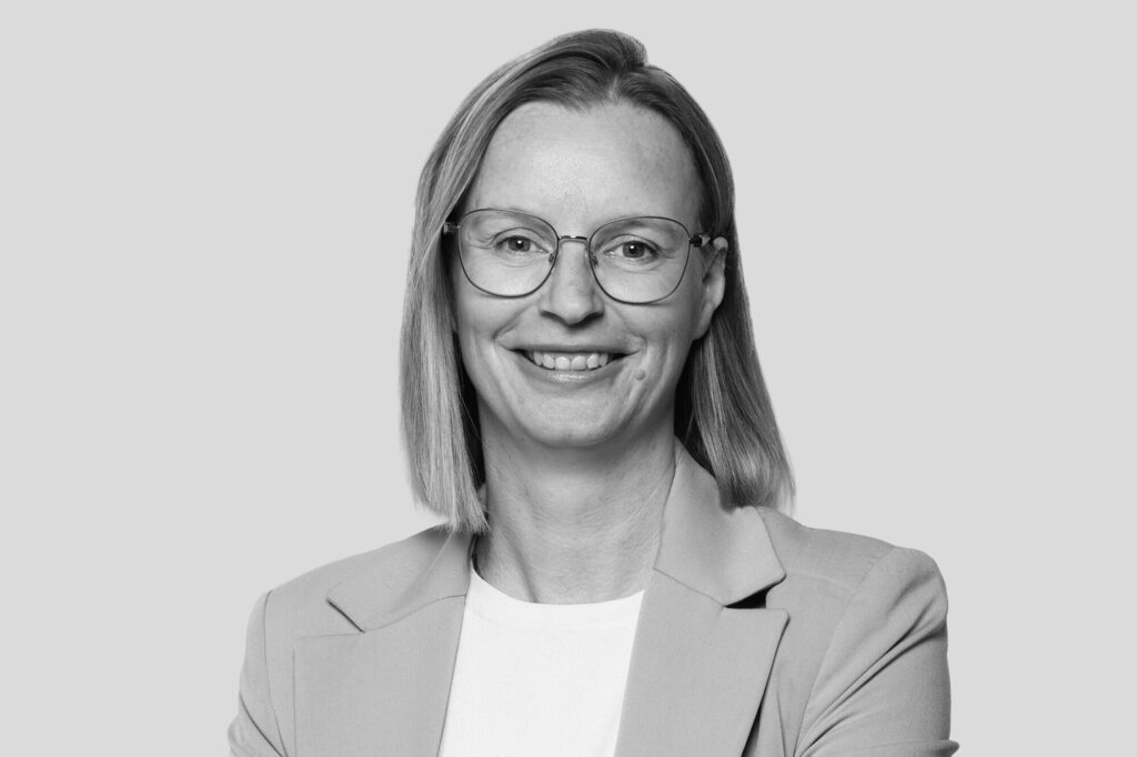 Porträt einer Frau mit Brille und schulterlangen Haaren, lächelnd, in Businesskleidung vor hellem Hintergrund.