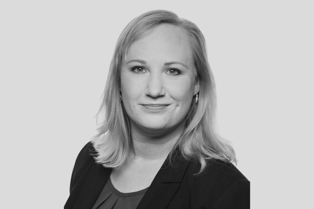 Porträt einer Frau mit schulterlangen Haaren, neutralem Gesichtsausdruck, in Businesskleidung vor hellem Hintergrund