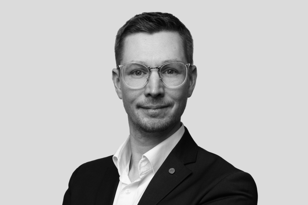Porträt eines Mannes in Businesskleidung vor neutralem Hintergrund. Er trägt ein dunkles Sakko über einem weißen Hemd und eine runde Brille. Das Bild ist in Schwarz-Weiß gehalten und wirkt professionell und ruhig.